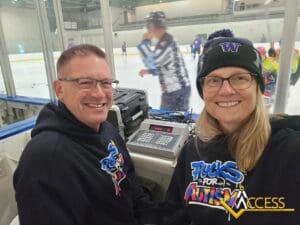 Two smiling people wearing Pucks for Autism hoodies sit in a hockey rink booth, with equipment in front of them. Players and a referee are visible on the ice in the background. A Access logo is at the bottom right.