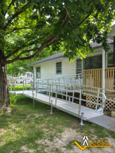 A metal wheelchair ramp leads from a grassy yard up to the porch of a light-colored house, shaded by a large tree. The ramp has railings and ends at a wooden deck. An ACCESS logo is in the bottom right corner.