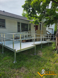 A silver metal wheelchair ramp with railings is installed outside a white house, leading up to the front porch. The yard is grassy with a large tree nearby. The Access logo is in the bottom right corner.