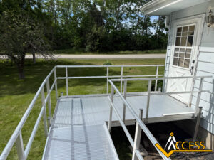 A metal wheelchair ramp with railings leads to a white door on a light blue house. Green grass, trees, and a road are visible in the background. The ramp has the Access company logo in the corner.