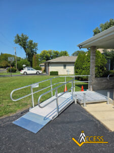 A metal wheelchair ramp with handrails leads from a sidewalk to a building entrance. Two orange cones are nearby. The ramp is on a paved area beside grass, with houses and trees in the background. ACCESS logo is in the corner.