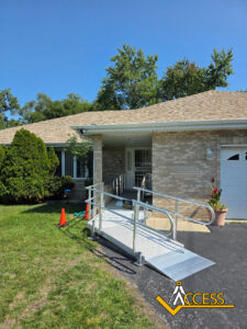 A silver wheelchair ramp with railings is installed leading to the front door of a brick house. Two orange cones are placed near the ramp, and the ACCESS logo appears in the bottom right corner.