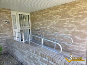 A metal wheelchair ramp with handrails is installed at the entrance of a brick house. The ramp leads to a white door. The logo ACCESS is in the bottom right corner.