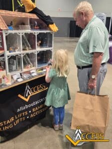 A young girl and an older man look at a detailed dollhouse with miniature stair lifts at an ACCESS booth. The man holds a paper bag while the girl explores the display. ACCESS branding is visible on the table.