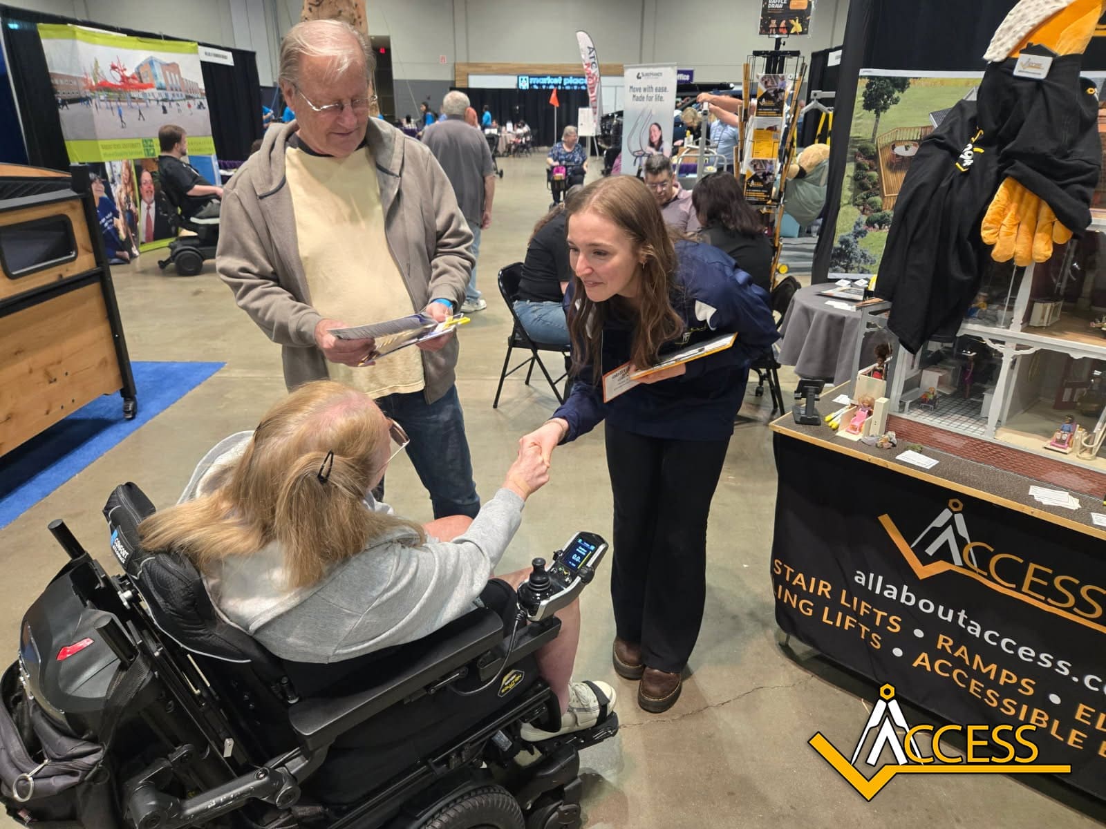 A woman in a wheelchair shakes hands with a smiling woman at an accessibility booth during an expo. A man stands nearby, holding papers. The booth displays brochures, ramps, and home accessibility products.