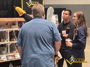 Three people converse at an expo booth featuring a large, detailed dollhouse display. Two individuals wear navy shirts with Access logos. Booth banners and an Access logo are visible in the background.