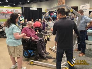 A man in a motorized wheelchair is being assisted by several people as he tries out a lift device at an indoor exhibition with various booths. The ACCESS logo is visible in the bottom right corner.