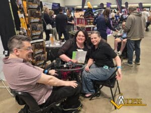 Three people, two using wheelchairs and one seated in a chair, smile together at an indoor expo. Booths, pamphlets, and crowds are visible in the background. A yellow Access logo appears in the corner.