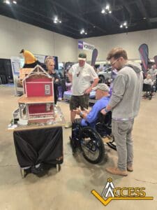 Four people gather around a detailed model of a red house at an indoor event. One man uses a wheelchair. Event booths are visible in the background, and an “Access” logo appears in the lower right corner.