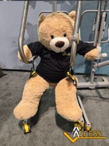 A large teddy bear wearing a black Access t-shirt sits secured in a mechanical lift on a gray carpet, with the Access company logo visible in the lower right corner.