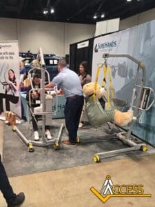 A man demonstrates a patient lift device with a woman at a trade show booth, while a large teddy bear rests in a sling attached to another lift. Booth banners and the Access logo are visible in the background.