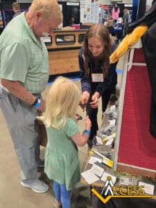 A young woman and an older man help a young blonde girl at a booth, showing her small items. The booth displays brochures and a model house, with the ACCESS logo visible in the lower right corner.