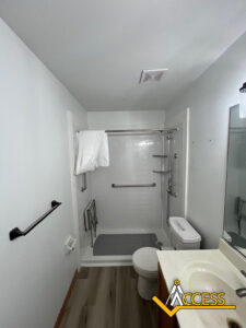 A small bathroom with a walk-in shower featuring grab bars and a fold-down seat, a toilet, a sink with a mirror, and light wood flooring. The ACCESS logo is in the bottom right corner.
