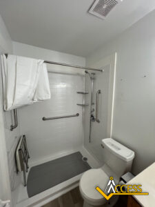 Accessible bathroom with a roll-in shower, fold-down seat, multiple grab bars, handheld showerhead, and a toilet. White tiles and fixtures, gray shower mat, and an “Access” logo in the bottom right corner.