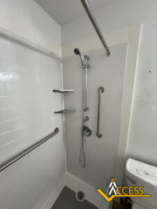 Accessible shower with white tile walls, a handheld showerhead, two grab bars, three corner shelves, a non-slip mat, and the ACCESS logo in the lower right corner. A toilet is partially visible on the right.
