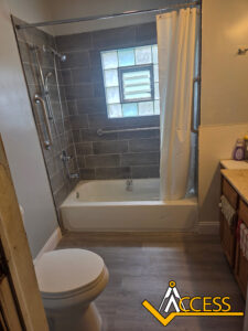 A bathroom with a bathtub and shower, grab bars installed on the wall, a toilet, a vanity with a sink, and a window above the tub. The ACCESS logo is visible in the bottom right corner.
