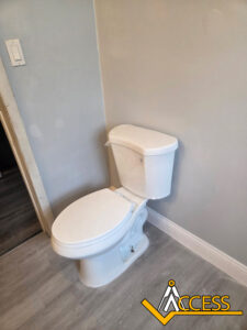 A clean, white toilet is positioned in the corner of a bathroom with light gray walls and wood-style flooring. The Access logo is in the bottom right corner.