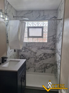 A modern bathroom with marble-patterned shower walls, a window with glass blocks, a grab bar, a white shower curtain, a black vanity with a sink, and the ACCESS logo in the bottom right corner.