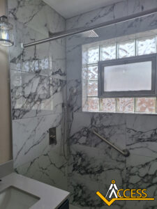 A modern bathroom shower with marble-patterned walls, a glass block window, a handheld showerhead, overhead rainfall shower, grab bar, and part of a sink. The Access company logo is visible in the bottom right corner.
