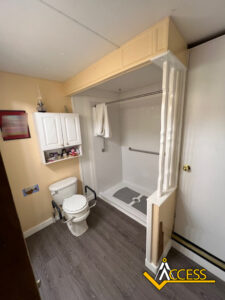 Accessible bathroom with a toilet equipped with grab bars, wall-mounted storage cabinets, and a step-in shower featuring a fold-down seat, grab bar, and handheld shower head. Access logo is visible in the corner.