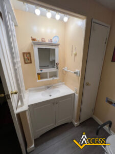 A small bathroom vanity with a sink, mirrored cabinet, wall-mounted shelves, and overhead lights. The walls are a soft peach color and there is a white door to the right. The ACCESS logo appears at the bottom right.