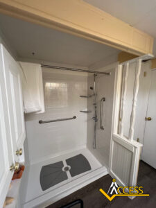 Accessible walk-in shower with grab bars, handheld showerhead, fold-down shower seat, and non-slip mat. White tile walls and curtain, partially open door, and Access logo in the bottom right corner.