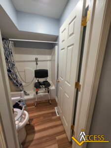 Small bathroom with an accessible shower area, featuring a shower chair, handheld showerhead, grab bar, shower curtain, and wood-style flooring. An open door partially covers the view. ACCESS logo in bottom right corner.