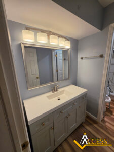 A modern bathroom with light blue walls, a white vanity with silver handles, a rectangular mirror, and a four-light fixture above. The ACCESS logo is in the bottom right corner.