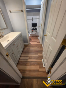 Accessible bathroom with wood-style flooring, a white vanity on the left, a grab bar on the wall, and a raised toilet seat with support handles in a small enclosed space. ACCESS logo in the bottom right corner.