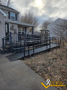 A black metal wheelchair ramp with railings leads from a driveway to the front porch of a beige house. The yard is bare, and the sky is overcast. The ACCESS logo is in the bottom right corner.