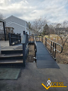 A black metal wheelchair ramp with handrails leads from a paved path to a porch next to stairs. Nearby houses and leafless trees are visible in the background. The Access logo appears in the bottom right corner.