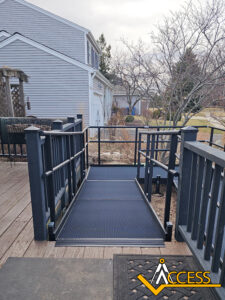A black metal wheelchair-accessible ramp with railings leads from a wooden deck to a paved walkway outside a gray house. The word “ACCESS” and a logo are in the bottom right corner.