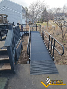 A black metal wheelchair ramp with railings leads from a porch to a paved path beside a house. The word ACCESS with a ramp icon is displayed in the bottom right corner.