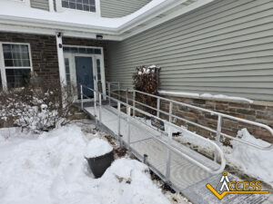 A metal wheelchair ramp with handrails leads to the entrance of a house in snowy conditions. Snow surrounds the ramp and bushes are nearby. An Access logo appears in the lower right corner.