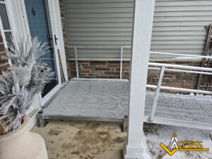 A metal wheelchair ramp with railings leads to a front door on a snowy day. The ramp is next to a house with brick and siding, and a winter planter is visible. The Access logo is in the bottom right corner.
