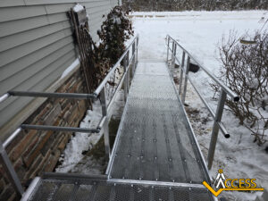 A metal wheelchair ramp with handrails leads from a snow-covered sidewalk to a house. Snow surrounds the area, and a bush and railing are visible beside the ramp. The ACCESS logo appears in the bottom right corner.
