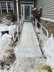 A metal wheelchair ramp with handrails leads to a front door of a house. The ground and ramp are surrounded by snow. There is a yellow Access logo in the bottom right corner.