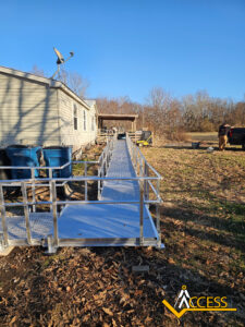 A long aluminum wheelchair ramp with handrails leads from a house to the yard. Two large blue barrels sit beside the house, and a person stands in the background near trees under a clear sky. Access company logo is in the corner.