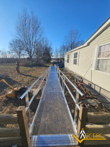 A silver metal wheelchair ramp extends from the entrance of a beige house to the yard, surrounded by bare trees and grass. The sky is clear and blue. A yellow ACCESS logo is in the bottom right corner.
