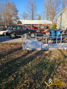 A silver wheelchair ramp leads from a grassy yard to a driveway with a black SUV, a blue garbage bin, and a carport with vehicles underneath. Leafless trees and a house are in the background. ACCESS logo is in the corner.