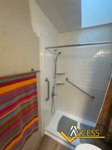 A walk-in shower with white tile walls, a handheld showerhead, grab bars, built-in shelves, and a bench. A colorful striped towel hangs outside. The Access company logo is in the lower right corner.
