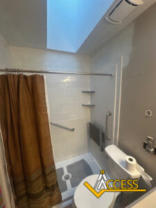 Small bathroom with a walk-in shower, white tile walls, brown shower curtain, grab bars, a fold-down bench, and a skylight above. Toilet with paper and air freshener nearby. ACCESS logo at bottom right.