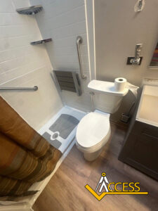 Accessible bathroom with a toilet next to a walk-in shower, equipped with grab bars, a fold-down shower seat, and a gray vanity. The floor is wood-style, and the Access company logo is visible.