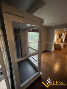 A residential indoor wheelchair lift is installed near a large window in a hallway with wooden floors. The Access company logo is visible in the lower right corner.