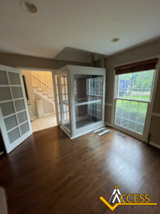 A residential room with hardwood floors features a partially installed home elevator near a set of stairs and a large window. French doors lean against one wall, and the “Access” logo is visible in the corner.