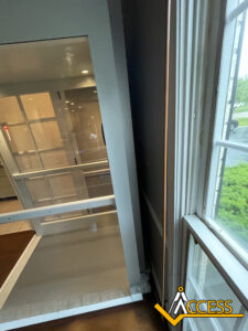 A view of an indoor wheelchair lift with glass panels, located next to a large window. The ACCESS logo is visible in the bottom-right corner.