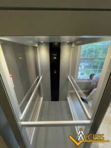 View from above of a modern residential elevator with glass sides, handrails, and control panel; a person is kneeling on the floor outside near a large window. The Access logo is in the bottom right corner.