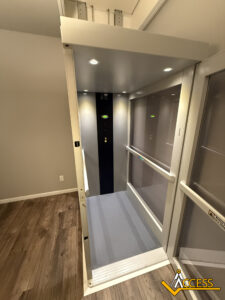 A modern indoor residential elevator with glass walls and a black control panel, illuminated by ceiling lights, located next to a wood-look floor; Access logo is visible in the bottom right corner.