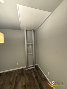 Minimalist room with gray walls and wood flooring featuring a retractable attic ladder partially extended from a ceiling hatch. A lamp is visible on the left, and the Access brand logo is in the bottom right corner.