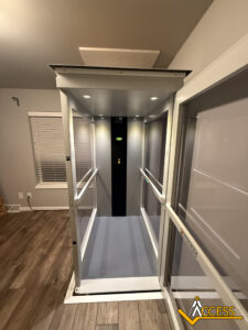 A modern residential elevator with glass walls, LED lighting, and a black control panel stands in a room with wood flooring and gray walls. The Access company logo is visible in the lower right corner.